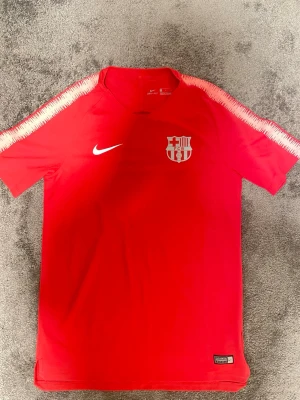 Röd FC Barcelona träningströja Nike - Snygg röd träningströja från Nike med FC Barcelona-logga på bröstet. Tröjan har vita detaljer längs axlarna och är tillverkad i ett lätt och snabbtorkande Dri-FIT-material. Perfekt för fotbollsträning eller gymmet.