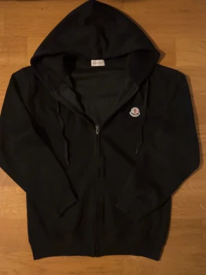Svart hoodie med dragkedja från Moncler - Säljer en svart hoodie från Moncler med dragkedja framtill och justerbar huva. Klassisk Moncler-logga på bröstet och ribbade muddar vid ärmslut och nederkant. Perfekt för dig som gillar stilrena och exklusiva plagg.