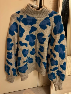 Grå stickad tröja med blått leopardmönster - Stickad tröja i grått med blått leopardmönster och hög ribbad polokrage. Tröjan har ballongärmar och breda muddar vid ärmslut och nederkant. 