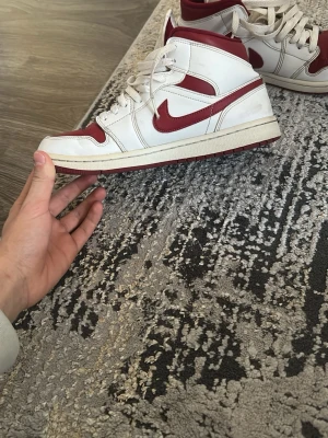 Nike Air Jordan 1 Mid röd/vit - Snygga Nike Air Jordan 1 Mid sneakers i vitt och rött läder. Klassisk siluett med röda detaljer och ikonisk Swoosh på sidan. Skorna har vita skosnören och en vit mellansula med röd yttersula. Perfekta för dig som gillar streetwear och vill sticka ut.