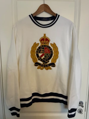 Vit sweatshirt från Polo Ralph Lauren - Limited edition säljs inte i butik längre!Vit sweatshirt med stort broderat emblem i guld, svart och rött på bröstet. Tröjan har svarta och vita ränder vid hals, ärmslut och nederkant. Klassisk och sportig stil med rund hals och långa ärmar.