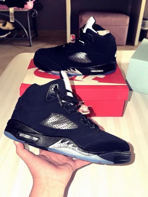 Air Jordan 5 Retro Black Metallic REFLECTIVE  - Snygga Air Jordan 5 Retro i svart med vita och röda detaljer. Skorna har en hög siluett, genomskinliga meshpaneler på sidorna och en isblå sula. Tungan är vit med röd Jumpman-logga och skorna har klassiska snörlås. Perfekta för dig som gillar streetwear och ikoniska sneakers.