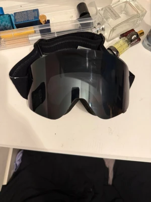 Svarta skidglasögon med spegellins - Säljer ett par svarta skidglasögon med bred, elastisk rem och spegellins. Glasögonen har mjuk skumvaddering på insidan för extra komfort och sitter stabilt på huvudet. Perfekta för skidåkning eller snowboard på vintern. Köpta för 2000kr använt bara en gång går inte att lämna tillbaks