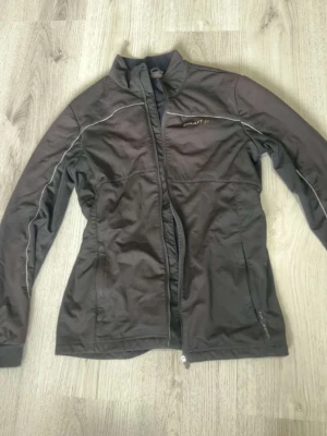 Svart träningsjacka från Craft - Sportig svart träningsjacka från Craft med hel dragkedja och diskreta vita detaljer längs sömmarna. Jackan har hög krage och är tillverkad i ett lätt, funktionellt material som passar perfekt för löpning eller utomhusträning.