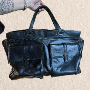 Svart handväska i läder med fickor - Stilren svart handväska i äkta läder med två stora fickor framtill och dubbla handtag. Väskan har en klassisk, fyrkantig form och detaljer i metall. Perfekt för dig som vill ha plats för allt och gillar en tidlös look.