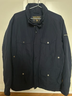 Marinblå vindjacka från Woolrich - Snygg marinblå vindjacka från Woolrich med fyra framfickor med lock och tryckknappar. Jackan har hög krage, dragkedja och knappslå samt grått innerfoder. Perfekt för vår- och höst dagar och stilren look. Den är helt ny.