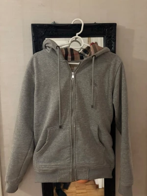 Grå hoodie med dragkedja från Burberry - Snygg grå hoodie med dragkedja från Burberry. Klassisk huva med dragsko och ikoniskt rutigt foder i beige, svart och rött. Mjukt material och två fickor framtill. Perfekt för en chill och stilren look.