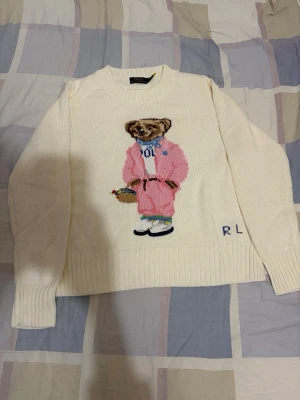 Ralph lauren björn stickat tröja - Vit stickad tröja från Polo Ralph Lauren med ett stort björnmotiv på framsidan där björnen bär rosa kläder. Tröjan har rund halsringning, ribbade muddar och RL-broderi nedtill. Perfekt för dig som gillar unika och lekfulla plagg.