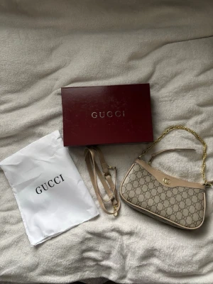 Beige GG Supreme handväska från Gucci - Säljer en beige GG Supreme handväska från Gucci med klassiskt monogrammönster, guldfärgad kedja och detaljer i ljusbrunt läder. Väskan har både axelrem och kedjehandtag, samt medföljande dustbag och originalkartong.