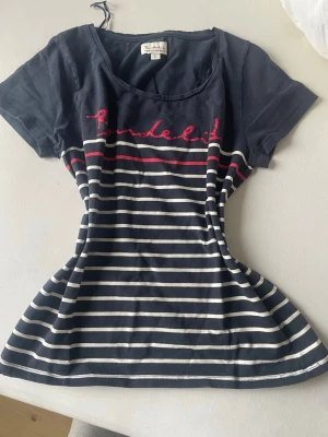 Randig t-shirt från Bondelid - Snygg marinblå t-shirt från Bondelid med vita och röda ränder samt röd broderad logga över bröstet. T-shirten har rund halsringning och korta ärmar. Perfekt för en avslappnad och stilren look.