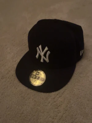Svart New Era Yankees keps - Snygg svart keps från New Era med klassiska New York Yankees-loggan broderad i vitt framtill. Modellen är 59FIFTY med platt skärm och sticker kvar på skärmen. Perfekt för dig som gillar streetwear och sportig stil.