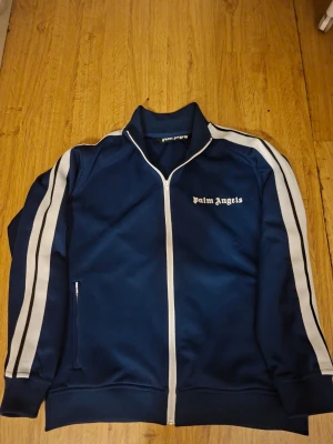 Blå track jacket från Palm Angels - Säljer en blå track jacket från Palm Angels med vita detaljer längs ärmarna och dragkedja framtill. Jackan har hög krage, vita ränder och logga på bröstet. Perfekt för en sportig och trendig look.