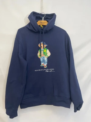 Ralph Lauren Bear hoodie  - Snygg mörkblå hoodie från Ralph Lauren med Polo Bear-tryck på bröstet. Klassisk huva med dragsko och ribbade muddar. Perfekt för dig som gillar streetwear och vill sticka ut med en ikonisk björn på tröjan.