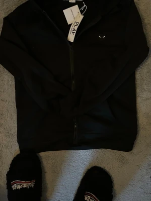 Svart hoodie från Comme des Garçons Play - Svart hoodie med dragkedja från Comme des Garçons Play. Klassisk design med huva och små vita ögonbroderier på bröstet. Materialet är mjukt och bekvämt, perfekt för en chill look. Taggar sitter kvar på plagget.