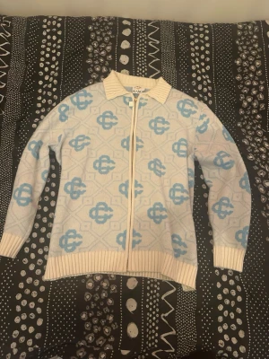 Vit och blå ziptröja från Casablanca - Säljer en vit stickad ziptröja från Casablanca med ljusblått mönster över hela tröjan. Tröjan har ribbade muddar, krage och dragkedja med snygg detalj. Perfekt för dig som gillar unik design och sköna material.