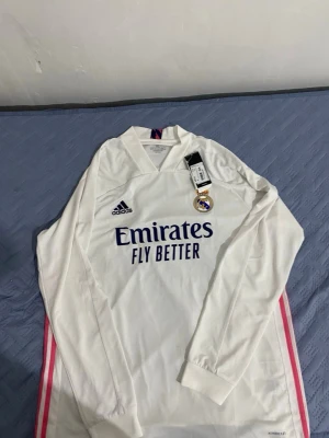 Vit långärmad Real Madrid-tröja Adidas - Säljer en vit långärmad Real Madrid-tröja från Adidas med Aeroready-material. Tröjan har klubbmärke på bröstet, Adidas-logga och sponsortryck i blått samt rosa detaljer längs sidorna. Perfekt för fotbollsträning eller att bära som supporter.