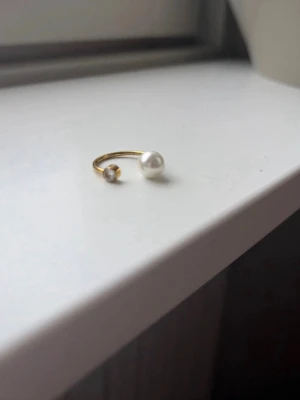 Guldfärgad öppen ring med pärlor - Trendig öppen ring i guldfärgad metall med två pärlor i olika storlekar, en vit och en grå. Minimalistisk och stilren design som ger en modern touch till din look.