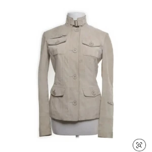 Beige mockajacka med ståkrage - Snygg beige mockajacka med ståkrage och spänne, fyra fickor med lock och knappar framtill. Jackan har en figurnära passform och långa ärmar med detaljspänne vid handlederna. Perfekt för dig som gillar stilrena och klassiska plagg.