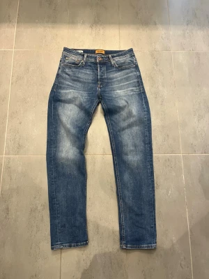 Blå jeans från Jack&jones - Säljer ett par klassiska blå jeans från jack&jones med raka ben. Jeansen har fem fickor, knappgylf och en läderpatch bak i midjan. Materialet är denim i bomull och färgen är mediumblå med ljusare partier.