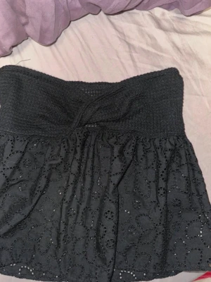 Svart broderad tubtopp med spetsdetaljer - Säljer en svart tubtopp med broderade blommönster och spetsdetaljer. Toppen har en twistad detalj framtill och är axelbandslös, vilket ger en snygg och somrig look. Perfekt att matcha med jeans eller kjol för en trendig outfit.