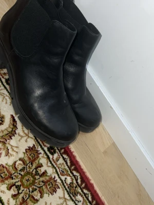 Svarta Chelsea boots i skinn - Säljer ett par klassiska svarta Chelsea boots i skinn med elastiska resårpaneler på sidorna och grov sula. Bootsens tidlösa design gör dem perfekta till många olika stilar. Klackhöjd och rundad tå ger en cool och bekväm look.