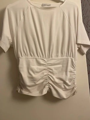 Vit topp från Anna Field - Trendig vit topp från Anna Field med korta ärmar och snygg smockdetalj framtill som ger en cool rynkad effekt. Tillverkad i mjukt material med rund halsringning. Perfekt för dig som vill ha en enkel men ändå unik topp.