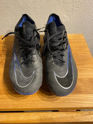 Nike Mercurial Zoom fotbollsskor svart/blå - Säljer ett par Nike Mercurial Zoom fotbollsskor i svart med blå detaljer och silvriga swoosh-loggor. Skorna har snörning, låg profil och ovandel i syntetmaterial för bra bollkontroll. Sulan är blå och designad för grepp på gräsplan. Perfekta för dig som vill ha speed och stil på planen.