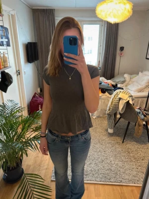 Mörkgrå peplum topp med kort ärm - Säljer en mörkgrå peplum topp med korta ärmar och rund hals. Toppen har en volang nedtill som ger en snygg och feminin siluett. Perfekt att matcha med jeans eller kjol för en trendig look.