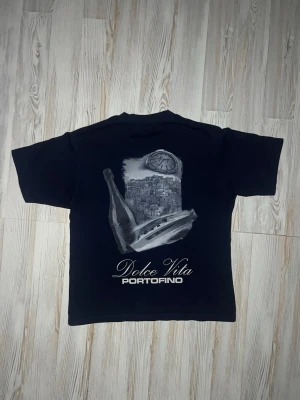 Svart Dolce Vita Portofino t-shirt - säljer denna marinblåa t-shirten med stor print på ryggen som visar en flaska, en klocka och en stadsvy🤩 Texten 'Dolce Vita PORTOFINO' är tryckt i vitt. Perfekt för dig som gillar unika motiv och italienska vibbar.😮‍💨