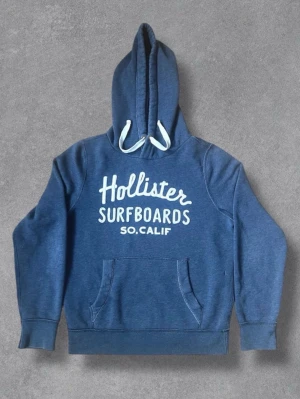 Blå hoodie från Hollister - Snygg blå hoodie från Hollister med vit text 'Hollister Surfboards SO.CALIF' på bröstet. Klassisk känguruficka framtill och vit dragsko i huvan. Perfekt för en avslappnad och cool stil.