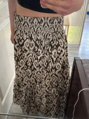 Mönstrad långkjol med resårmidja - Säljer en svart och beige långkjol med resår i midjan och ett coolt abstrakt mönster. Kjolen är luftig och har en avslappnad passform, perfekt för varma dagar. Materialet känns mjukt och skönt mot huden.