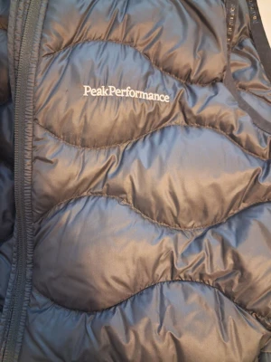 Svart dunväst från Peak Performance - Snygg svart dunväst från Peak Performance med quiltat mönster och dragkedja framtill. Västen har hög krage och är tillverkad i glansigt material med diskret logga på bröstet. Perfekt för lager-på-lager-stil under kyliga dagar.