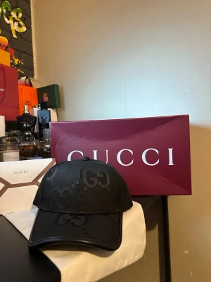 Svart keps från Gucci med GG-mönster - Snygg svart keps från Gucci med klassiskt GG-monogram i ton-i-ton. Kepsen har böjd skärm och justerbar baksida. Tillverkad i ett mesh-liknande material som ger en sportig och exklusiv känsla. Perfekt accessoar för att lyfta din outfit.