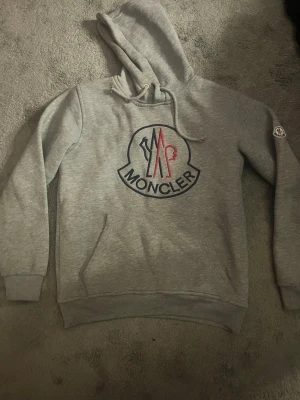 Grå Moncler hoodie med logga - Snygg grå hoodie från Moncler med stor logga i blått och rött på bröstet. Klassisk huva med dragsko och ribbade muddar. Materialet är mjukt och bekvämt, perfekt för en chill look. Moncler-märke på vänster ärm. Känguruficka framtill. Kom privat vid frågor eller flera bilder👍🏼