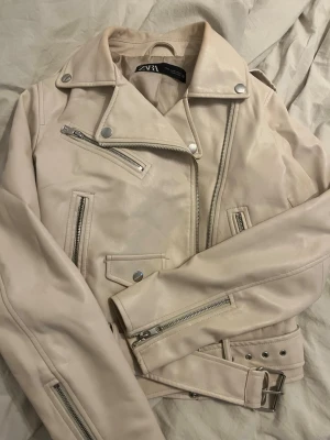 Ljusbeige skinnjacka från Zara - Snygg ljusbeige skinnjacka från Zara med klassisk bikerstil. Jackan har flera silvriga dragkedjor, axelklaffar och bälte i midjan. Perfekt för dig som vill ha en stilren och trendig look med coola detaljer.