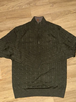 Mörkgrön stickad tröja Morris - Mörkgrön stickad tröja från Morris med kabelstickat mönster och halv dragkedja framtill. Tröjan har hög krage med kontrastfärg på insidan och en liten broderad logga på bröstet. Perfekt för dig som gillar klassisk men avslappnad stil.