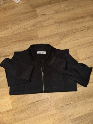 Mörk grå overshirt från Samsøe Samsøe - Stilren mörk grå overshirt från Samsøe Samsøe med klassisk krage, dragkedja framtill och bröstficka. Overshirten är  i ett mjukt tyg som ger en avslappnad look. Perfekt att bära över en t-shirt eller hoodie.