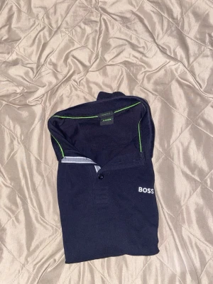 Svart pikétröja från BOSS - Klassisk mörkblå pikétröja från BOSS med vit logga på bröstet. Tröjan har korta ärmar, knappslå och kontrastdetaljer i vitt och grönt på kragen och ärmsluten. Perfekt till en stilren och avslappnad look.