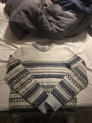 Grå stickad tröja med mönster - Säljer en grå stickad tröja med mörkblå och vita detaljer i ett klassiskt nordiskt mönster. Tröjan har rund halsringning. Perfekt för kalla dagar och riktigt snygg till jeans.