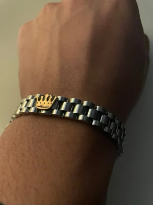 R@lex liknade armband  - Snyggt armband i silverfärgad metall med länkdesign och en guldfärgad krona i mitten. Armbandet har en stilren och modern look som passar till många olika stilar. Perfekt accessoar för dig som gillar detaljer med lite edge.