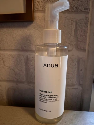 Anua Heartleaf Cleansing Oil - Anua Heartleaf Pore Control Cleansing Oil i genomskinlig plastflaska med vit pump. Oljan är framtagen för att rengöra porer och ta bort smink. Enkel och stilren design med vit etikett och svart text.