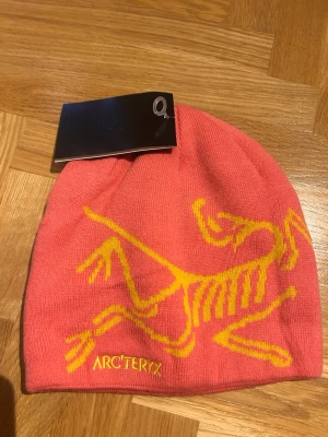 Aprikosfärgad mössa från Arc'teryx - Säljer en snygg rosa mössa från Arc'teryx med gult skelettmönster och broderad logga framtill. Mössan är stickad och har en mjuk, följsam passform. Perfekt för dig som vill ha något färgglatt och sportigt till vintern.