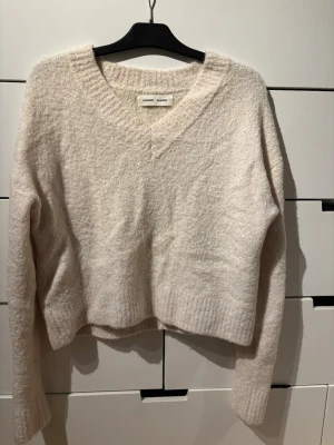 Beige stickad tröja Samsoe Samsoe - Mysig beige stickad tröja från Samsoe Samsoe med v-ringning och lång ärm. Tröjan har en lite boxig, croppad passform och är gjord i ett mjukt, fluffigt material. Perfekt för lager på lager och enkel att matcha till olika outfits.