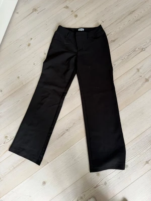 Svarta kostymbyxor med raka ben - Snygga svarta kostymbyxor med raka ben och klassisk passform. Byxorna har hällor för skärp och stängs med dragkedja och knapp. Perfekta för en clean och stilren look. Materialet ser ut att vara en mjuk och lite stretchig bomullsblandning.