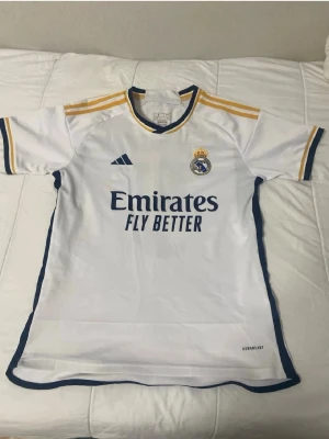 Real Madrid matchtröja Adidas vit - Säljer en vit Real Madrid matchtröja från Adidas med gula och mörkblå detaljer. Tröjan har klubbmärket på bröstet, Adidas-logga och sponsortryck 'Emirates Fly Better' framtill. Tillverkad i lätt och ventilerande material, perfekt för fotbollsträning eller match.