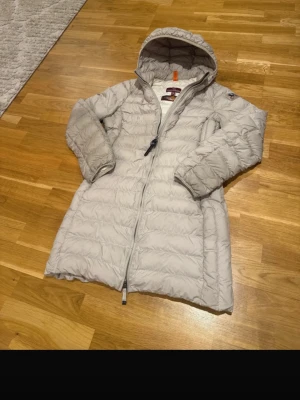 Ljusgrå dunjacka från Parajumpers - Snygg ljusgrå dunjacka från Parajumpers med huva och dragkedja framtill. Jackan är lättviktig och har en längre modell som ger extra värme. Perfekt för kalla vinterdagar och har en diskret logga på ärmen.