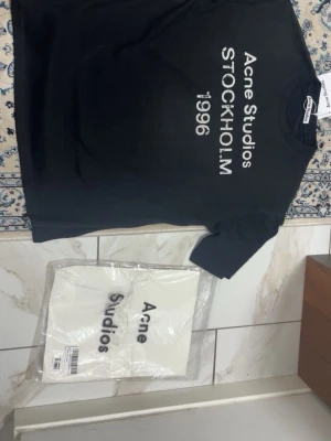 Acne Studios T-Shirt - Svart t-shirt från Acne Studios med distressed texttryck. Helt ny, papperstagen avklippt men sitter kvar i nedre delen av t-shirten. Stilren modell i mjuk och högkvalitativ bomull.