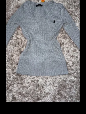 Grå stickad tröja från Ralph Lauren - Supersnygg grå stickad tröja från Ralph Lauren med smal passform och v-ringning. Tröjan har långa ärmar och klassisk ribbstickning samt den ikoniska svarta loggan broderad på bröstet. Perfekt till jeans eller kjol.