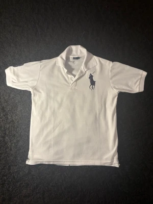 Vit pikétröja från Ralph Lauren - Klassisk vit pikétröja från Ralph Lauren med kort ärm och stor mörkblå broderad logga på bröstet. Tröjan har krage och knäppning framtill. Perfekt för en clean och sportig stil.