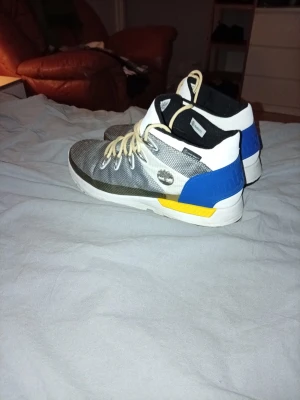 Färgblockade Timberland sneakers - Snygga sneakers från Timberland med grå mesh framtill, vita och svarta detaljer, samt blå och gula färgblock på hälen. Storlek 43. Köpte dom i november. Mycket bra skick. Nytt pris 1400 mitt pris 500